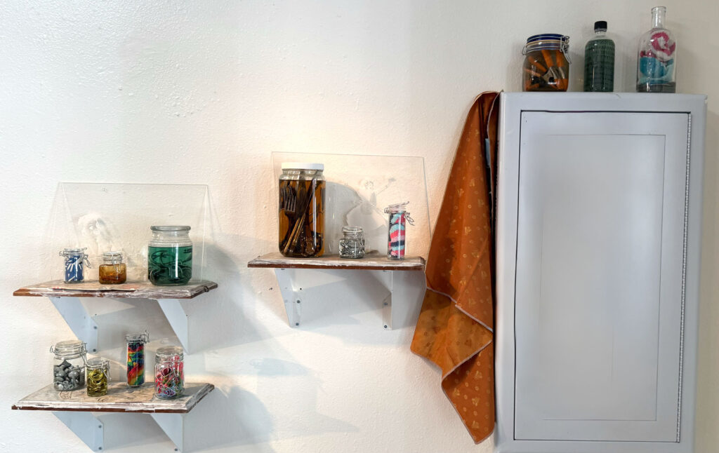 wall installation with glass jars containing harware, cutlery, yarn, fabric, collaged shelves