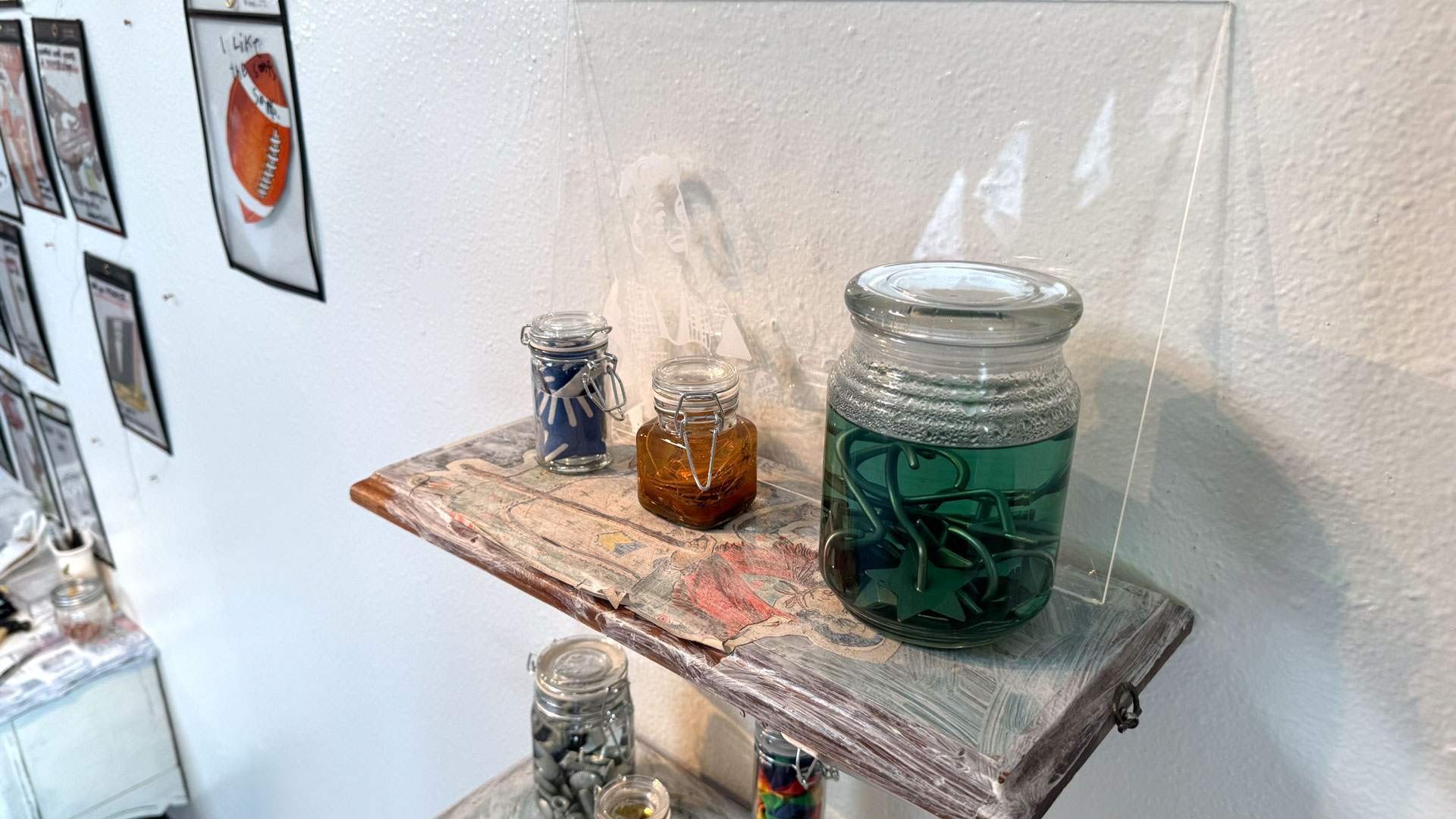 wall installation with glass jars containing harware, cutlery, yarn, fabric, collaged shelves